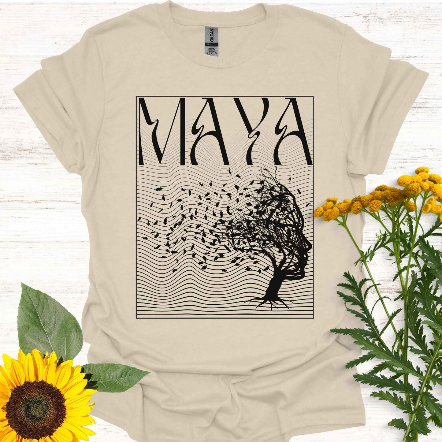 Beige t-shirt featuring the word 'Maya' above an optical illusion graphic of a tree shaped like a face, with leaves blowing in the wind and an abstract wavy background pattern. The design symbolizes the Sanskrit concept of 'maya,' meaning illusion, highlighting the illusory nature of reality shaped by the mind. It evokes themes of duality, self-reflection, and the power of thought in creating our perceptions, encouraging deeper awareness and questioning of assumptions about reality.