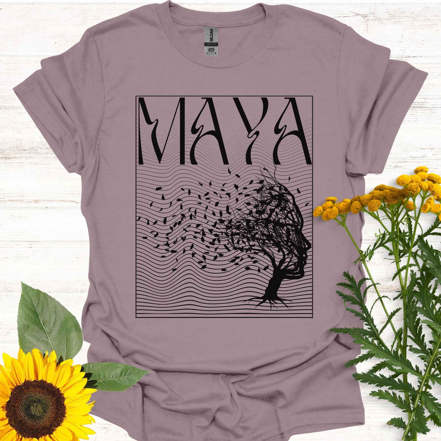 Light purple t-shirt featuring the word 'Maya' above an optical illusion graphic of a tree shaped like a face, with leaves blowing in the wind and an abstract wavy background pattern. The design symbolizes the Sanskrit concept of 'maya,' meaning illusion, highlighting the illusory nature of reality shaped by the mind. It evokes themes of duality, self-reflection, and the power of thought in creating our perceptions, encouraging deeper awareness and questioning of assumptions about reality.