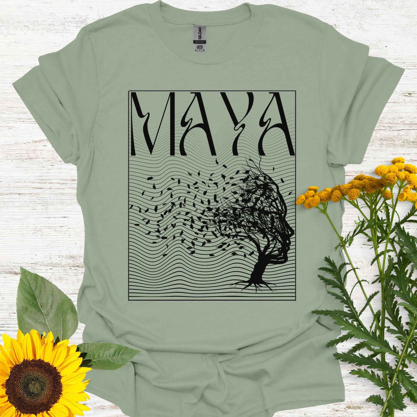 Sage green t-shirt featuring the word 'Maya' above an optical illusion graphic of a tree shaped like a face, with leaves blowing in the wind and an abstract wavy background pattern. The design symbolizes the Sanskrit concept of 'maya,' meaning illusion, highlighting the illusory nature of reality shaped by the mind. It evokes themes of duality, self-reflection, and the power of thought in creating our perceptions, encouraging deeper awareness and questioning of assumptions about reality.
