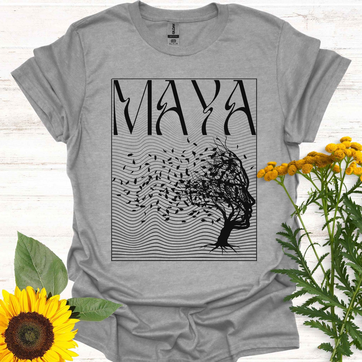 Grey t-shirt featuring the word 'Maya' above an optical illusion graphic of a tree shaped like a face, with leaves blowing in the wind and an abstract wavy background pattern. The design symbolizes the Sanskrit concept of 'maya,' meaning illusion, highlighting the illusory nature of reality shaped by the mind. It evokes themes of duality, self-reflection, and the power of thought in creating our perceptions, encouraging deeper awareness and questioning of assumptions about reality.