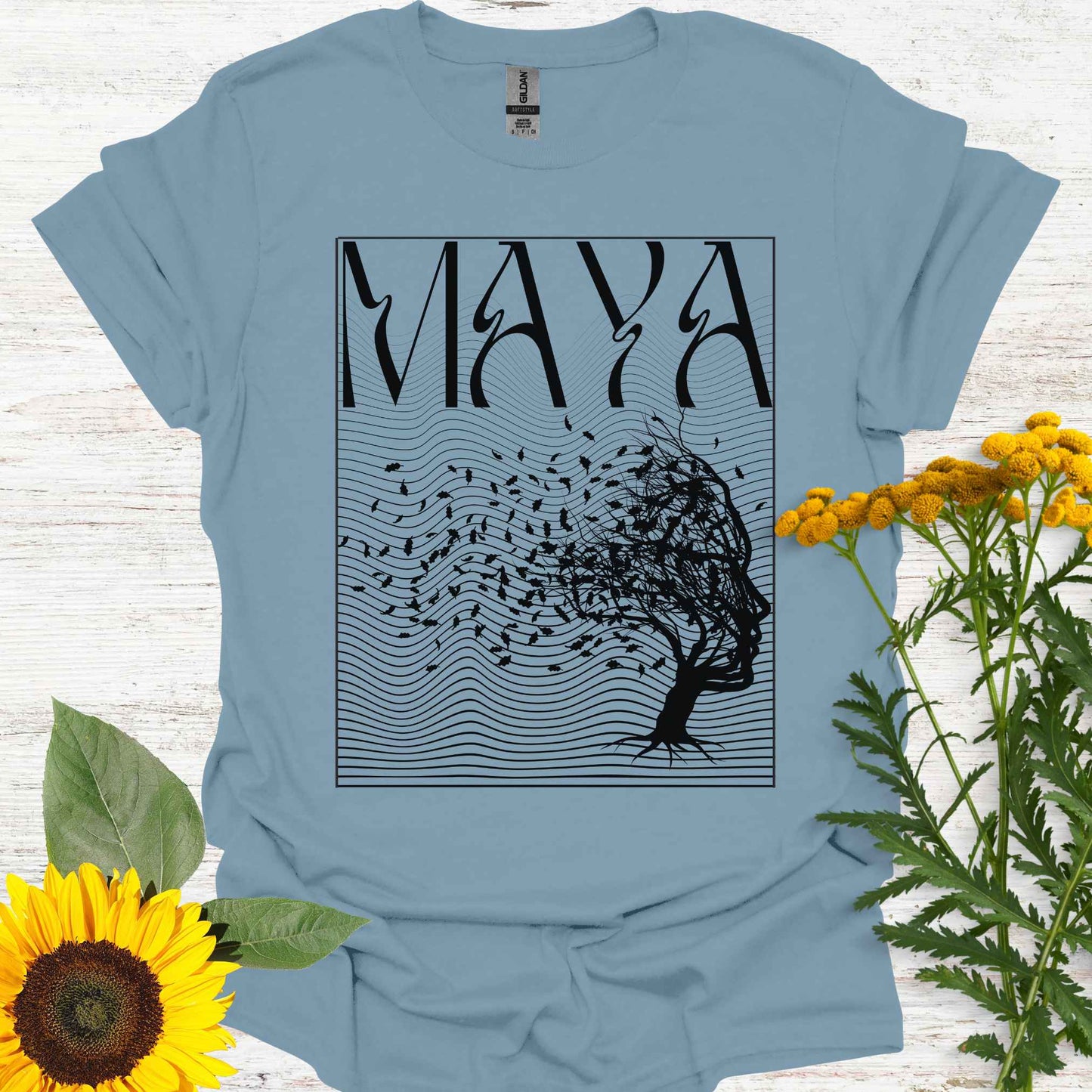 Blue t-shirt featuring the word 'Maya' above an optical illusion graphic of a tree shaped like a face, with leaves blowing in the wind and an abstract wavy background pattern. The design symbolizes the Sanskrit concept of 'maya,' meaning illusion, highlighting the illusory nature of reality shaped by the mind. It evokes themes of duality, self-reflection, and the power of thought in creating our perceptions, encouraging deeper awareness and questioning of assumptions about reality.