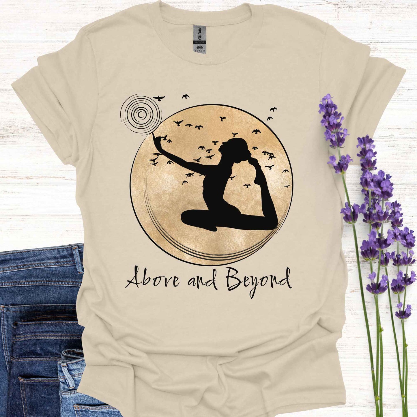 Beige t-shirt design featuring a design with a female figure in a yoga pose, one hand pointing upwards, set against a framed gold circular background with birds in flight. Below, the phrase 'Above and Beyond' signifies transcending limitations and striving for personal growth. Ideal for yoga enthusiasts and those on a journey of self-improvement.