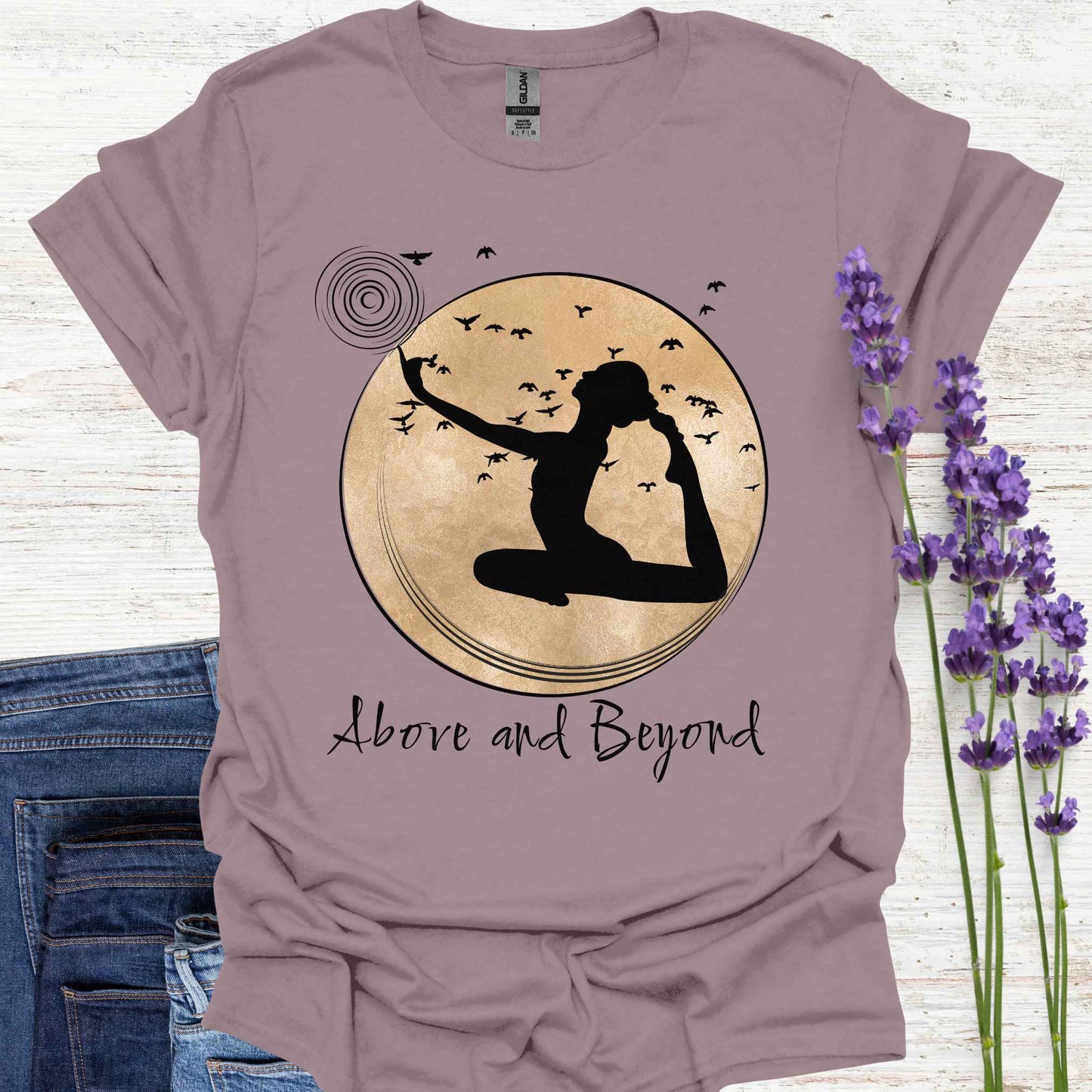 Light purple t-shirt design featuring a design with a female figure in a yoga pose, one hand pointing upwards, set against a framed gold circular background with birds in flight. Below, the phrase 'Above and Beyond' signifies transcending limitations and striving for personal growth. Ideal for yoga enthusiasts and those on a journey of self-improvement.