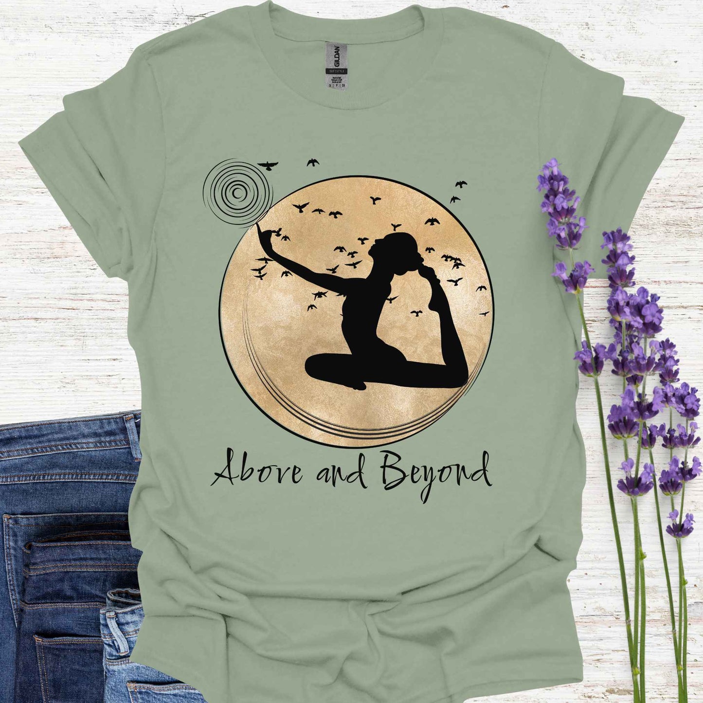 Sage green t-shirt design featuring a design with a female figure in a yoga pose, one hand pointing upwards, set against a framed gold circular background with birds in flight. Below, the phrase 'Above and Beyond' signifies transcending limitations and striving for personal growth. Ideal for yoga enthusiasts and those on a journey of self-improvement.