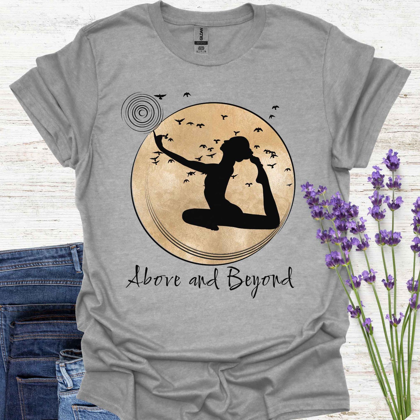 White t-shirt design featuring a design with a female figure in a yoga pose, one hand pointing upwards, set against a framed gold circular background with birds in flight. Below, the phrase 'Above and Beyond' signifies transcending limitations and striving for personal growth. Ideal for yoga enthusiasts and those on a journey of self-improvement.
