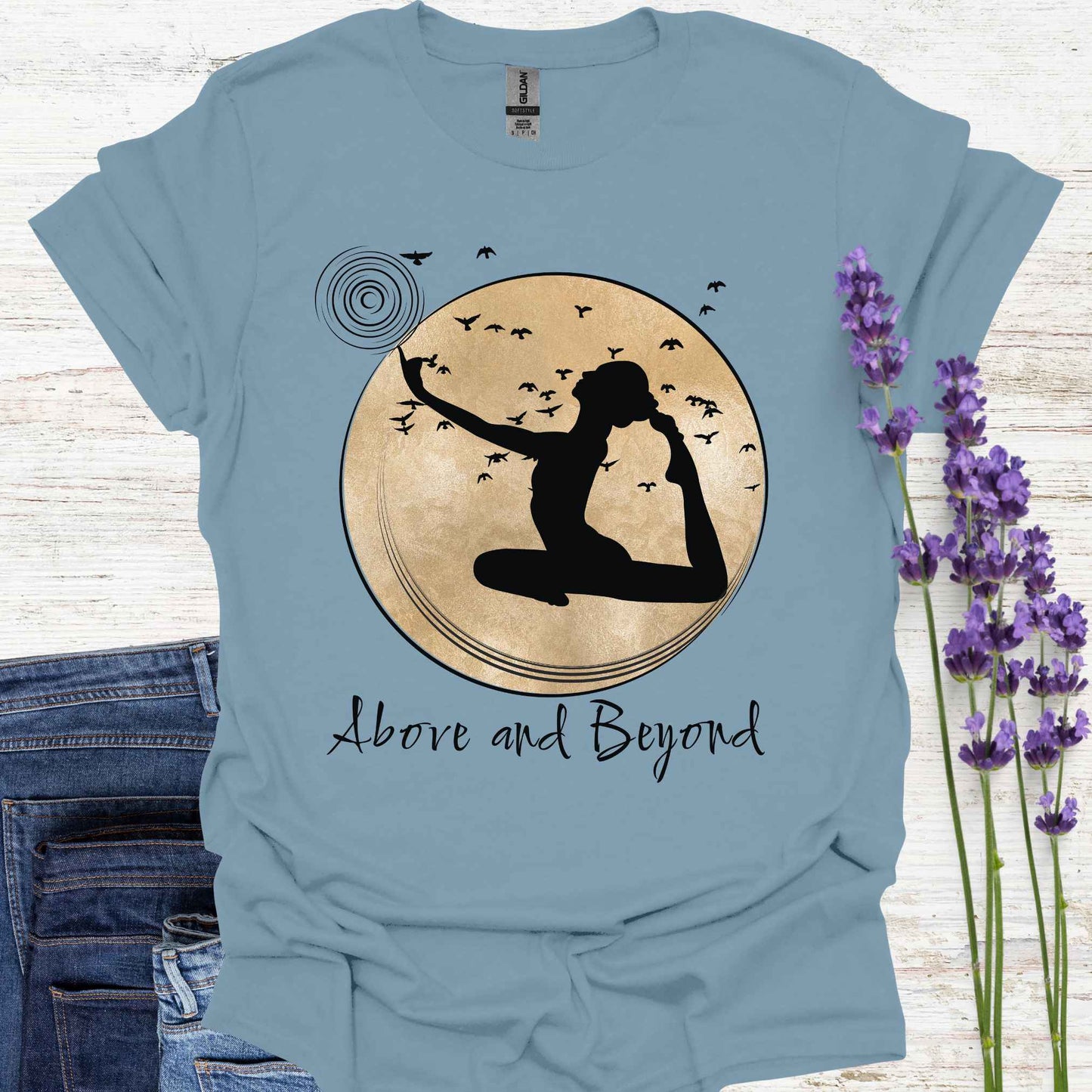Blue t-shirt design featuring a design with a female figure in a yoga pose, one hand pointing upwards, set against a framed gold circular background with birds in flight. Below, the phrase 'Above and Beyond' signifies transcending limitations and striving for personal growth. Ideal for yoga enthusiasts and those on a journey of self-improvement.
