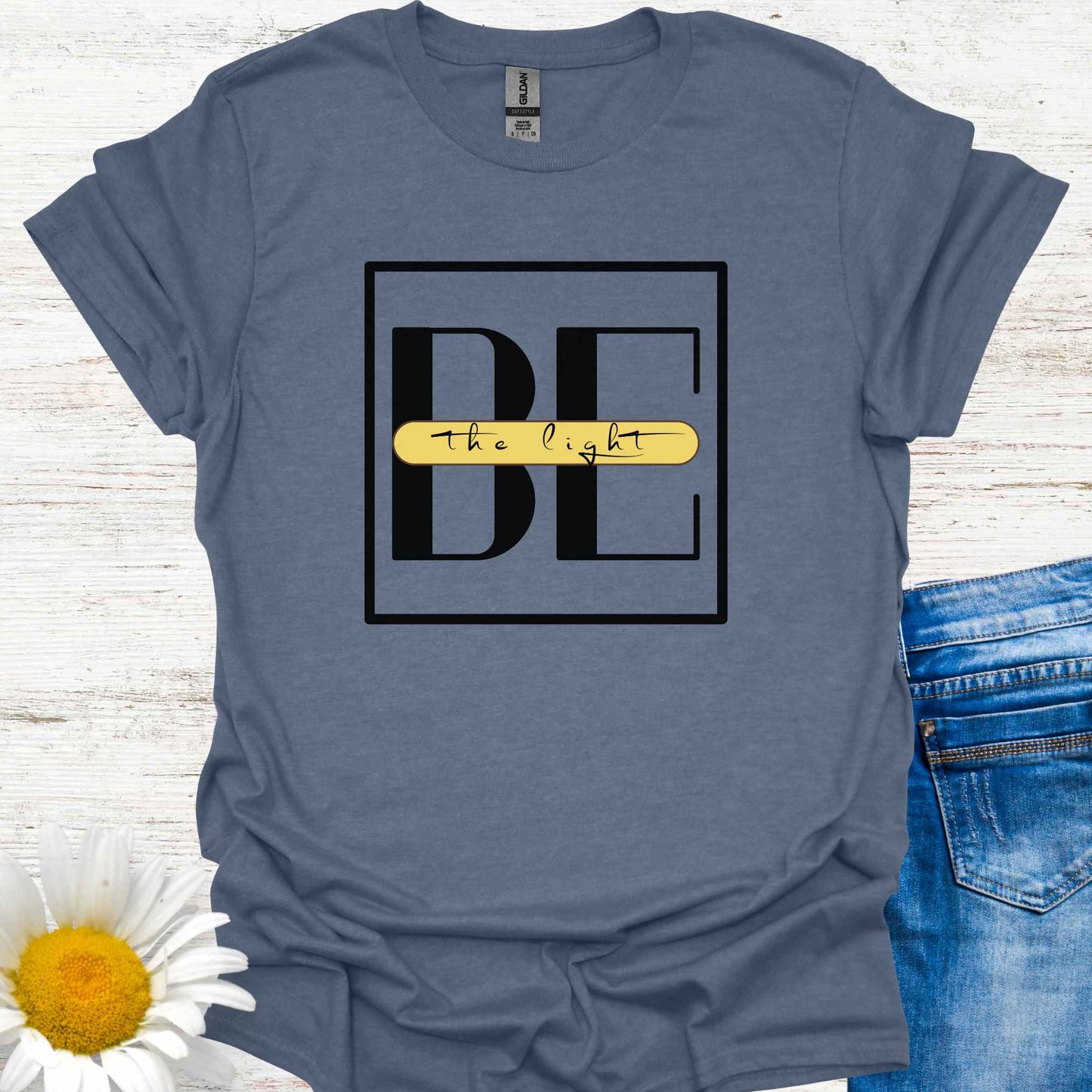 Blue t-shirt featuring the phrase 'Be the Light,' with the word 'BE' in large letters and the small cursive words 'the light' written across it on a yellow banner. This design encourages positivity, inspiration, and personal empowerment, reminding wearers to shine their light and spread kindness in the world.