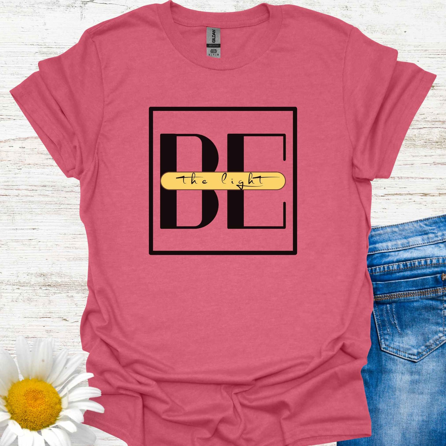 Red t-shirt featuring the phrase 'Be the Light,' with the word 'BE' in large letters and the small cursive words 'the light' written across it on a yellow banner. This design encourages positivity, inspiration, and personal empowerment, reminding wearers to shine their light and spread kindness in the world.