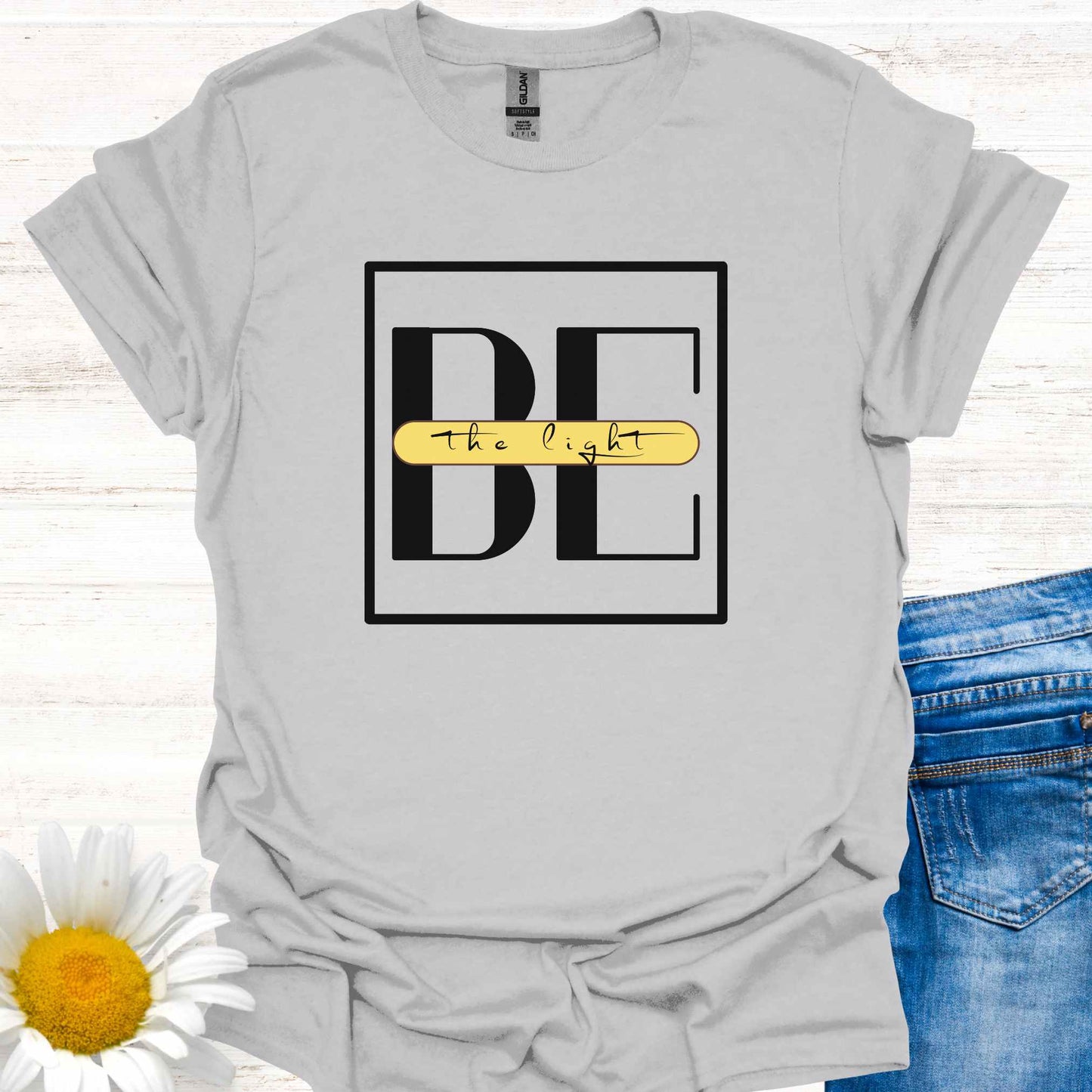 Grey t-shirt featuring the phrase 'Be the Light,' with the word 'BE' in large letters and the small cursive words 'the light' written across it on a yellow banner. This design encourages positivity, inspiration, and personal empowerment, reminding wearers to shine their light and spread kindness in the world.