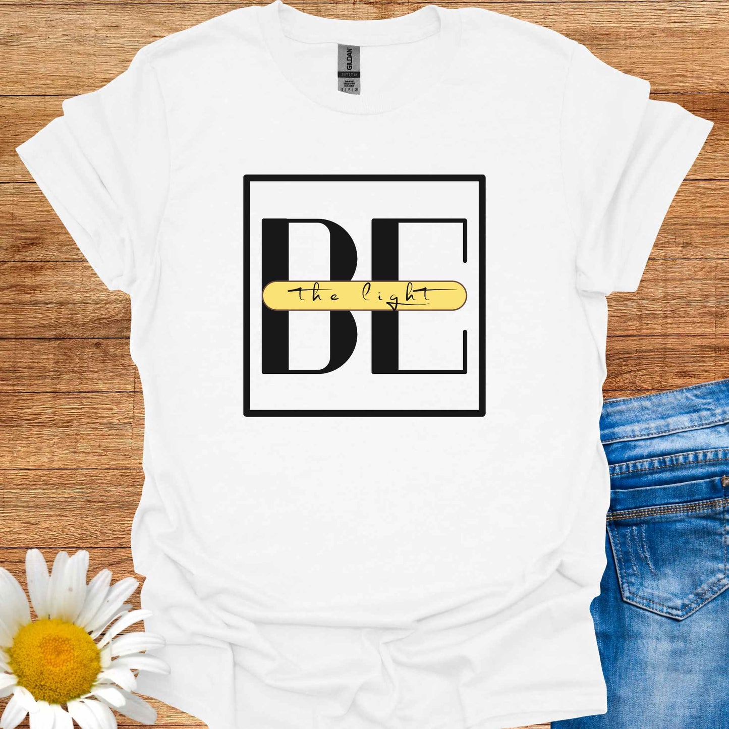 White t-shirt featuring the phrase 'Be the Light,' with the word 'BE' in large letters and the small cursive words 'the light' written across it on a yellow banner. This design encourages positivity, inspiration, and personal empowerment, reminding wearers to shine their light and spread kindness in the world.