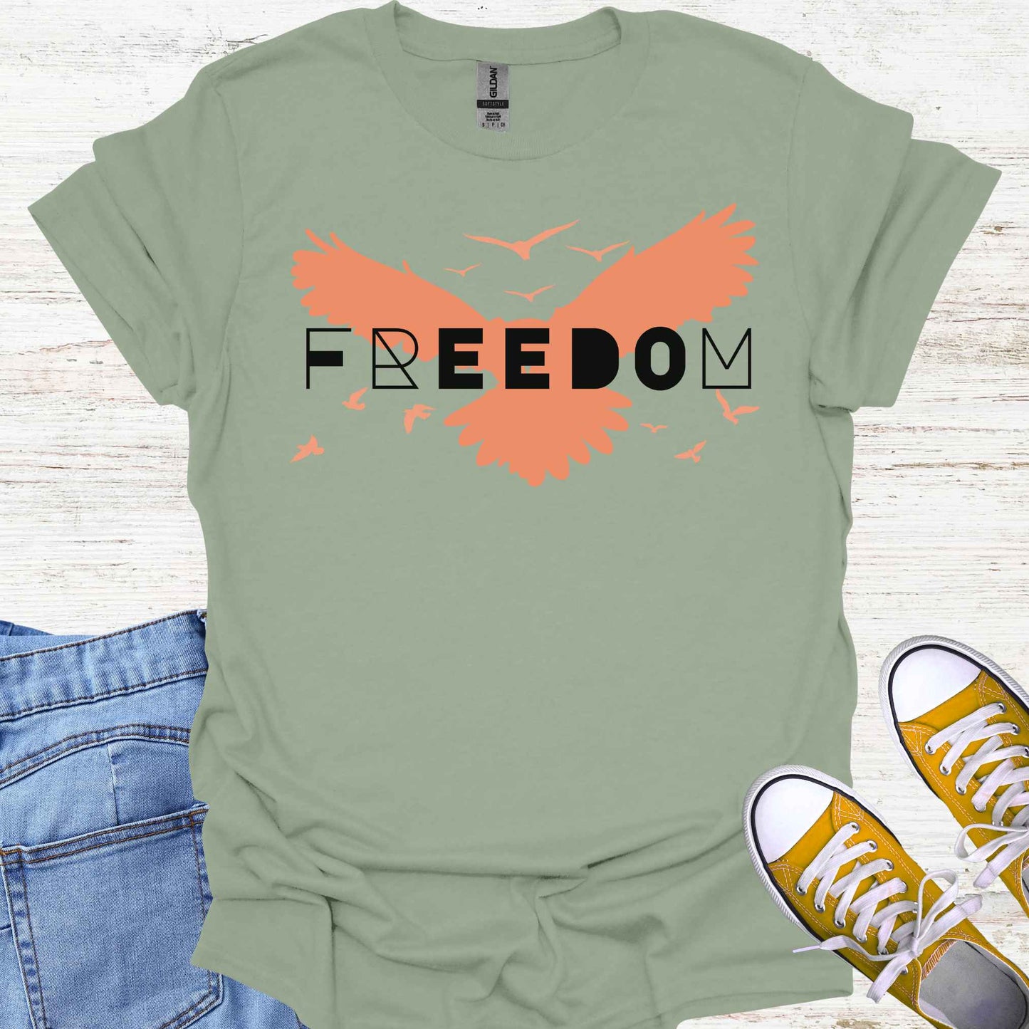 Sage green t-shirt featuring an orange bird with its wings spread, symbolizing freedom and liberation, with the bold word 'FREEDOM' in stylish typography placed on top. This design evokes the spirit of independence, self-expression, and the boundless potential of the human soul, inspired by the free flight of birds.