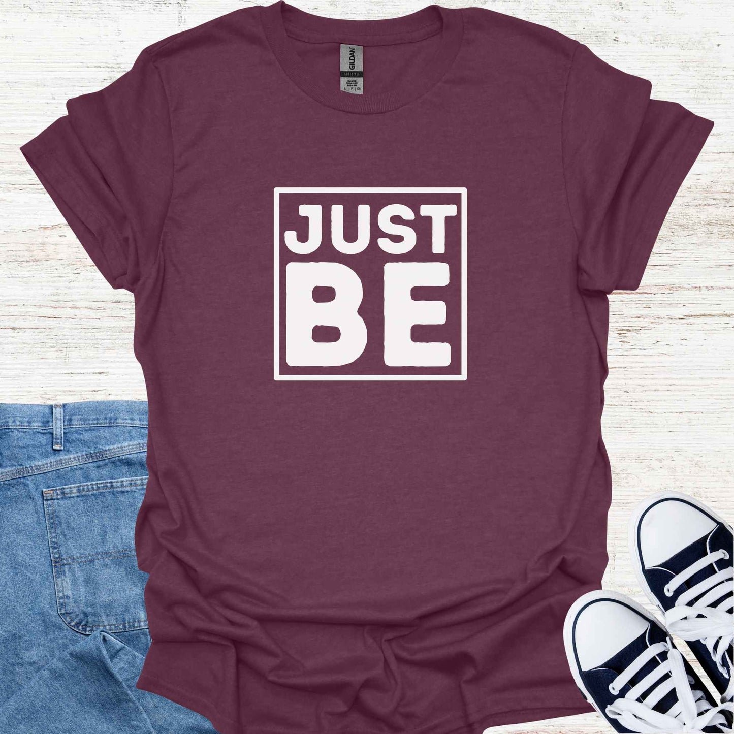 Purple t-shirt design featuring the minimalist phrase 'Just Be' in bold typography, framed for emphasis. The design conveys a profound spiritual message about living in the present moment, embracing simplicity, and finding peace in existence itself. It encourages mindfulness, acceptance, and the art of simply being, free from the need to do or become anything. Perfect for those drawn to spirituality, introspection, and mindful living.