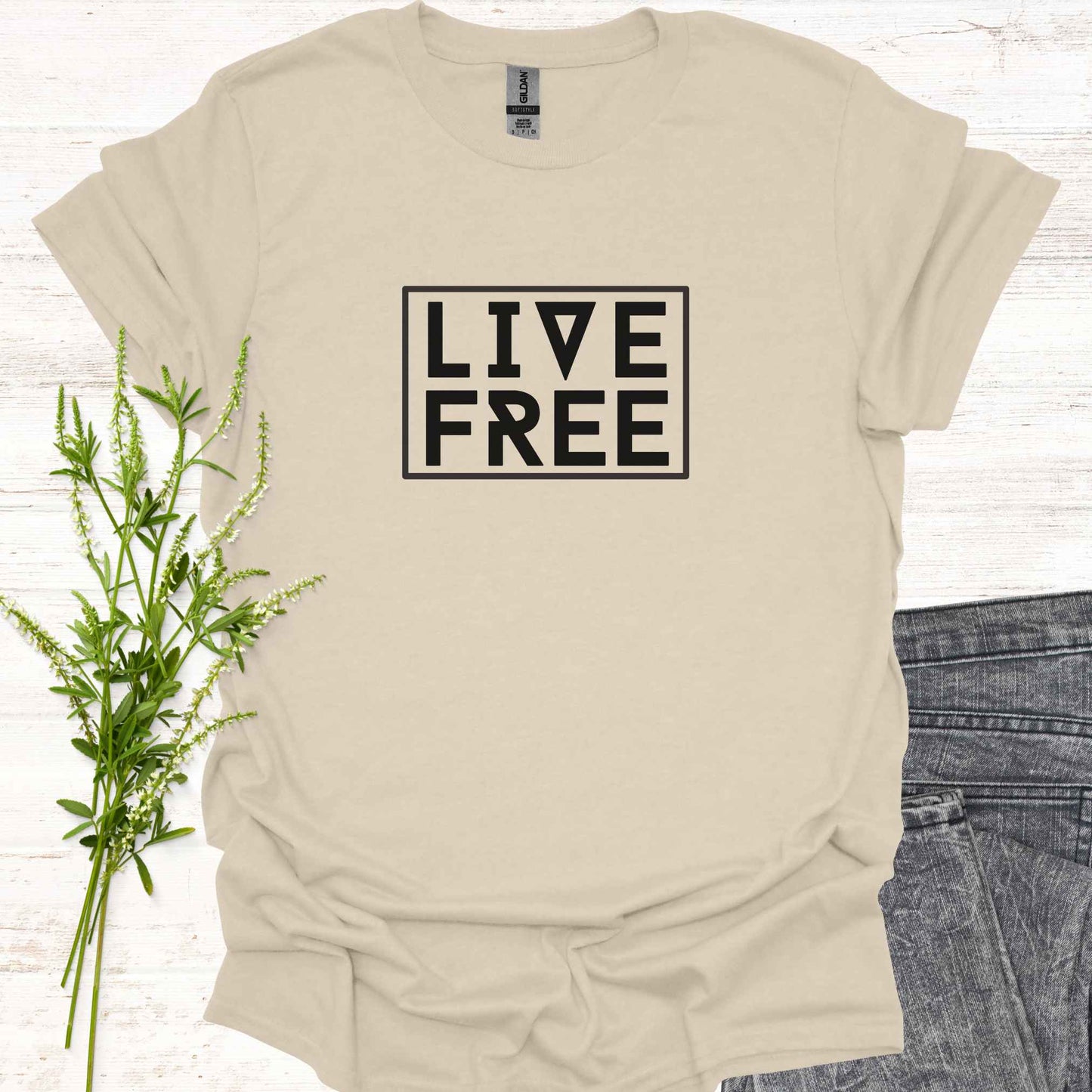 Beige t-shirt featuring bold text-based design with the phrase 'Live Free' framed on a minimalist background. This design embodies the spirit of independence, self-determination, and breaking free from mental constraints. It encourages a life aligned with personal values and freedom of thought, resonating with themes of spiritual liberation, individuality, and self-empowerment.
