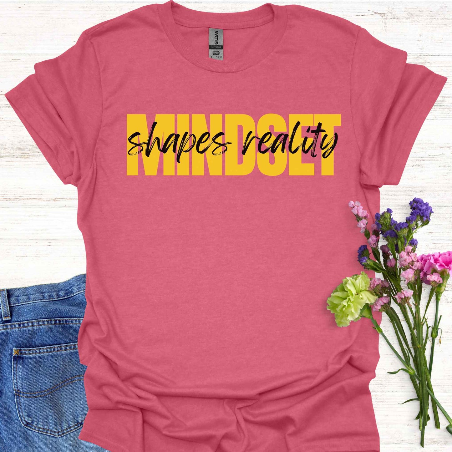 Red t-shirt featuing a text-based design with the phrase 'Mindset Shapes Reality'. The word 'Mindset' is in bold yellow capital letters, while 'shapes reality' is in smaller cursive lettering. This design highlights how our mindset influences our perception and reality, empowering us to take control of our experiences and perspective in life.