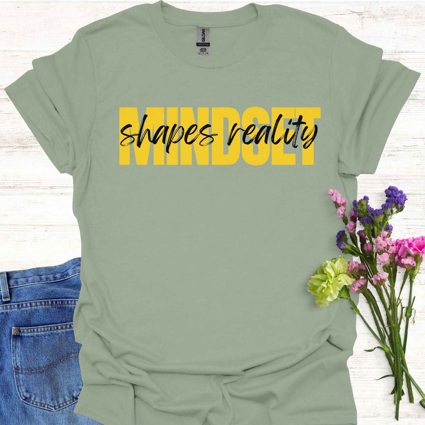 Sage green t-shirt featuing a text-based design with the phrase 'Mindset Shapes Reality'. The word 'Mindset' is in bold yellow capital letters, while 'shapes reality' is in smaller cursive lettering. This design highlights how our mindset influences our perception and reality, empowering us to take control of our experiences and perspective in life.