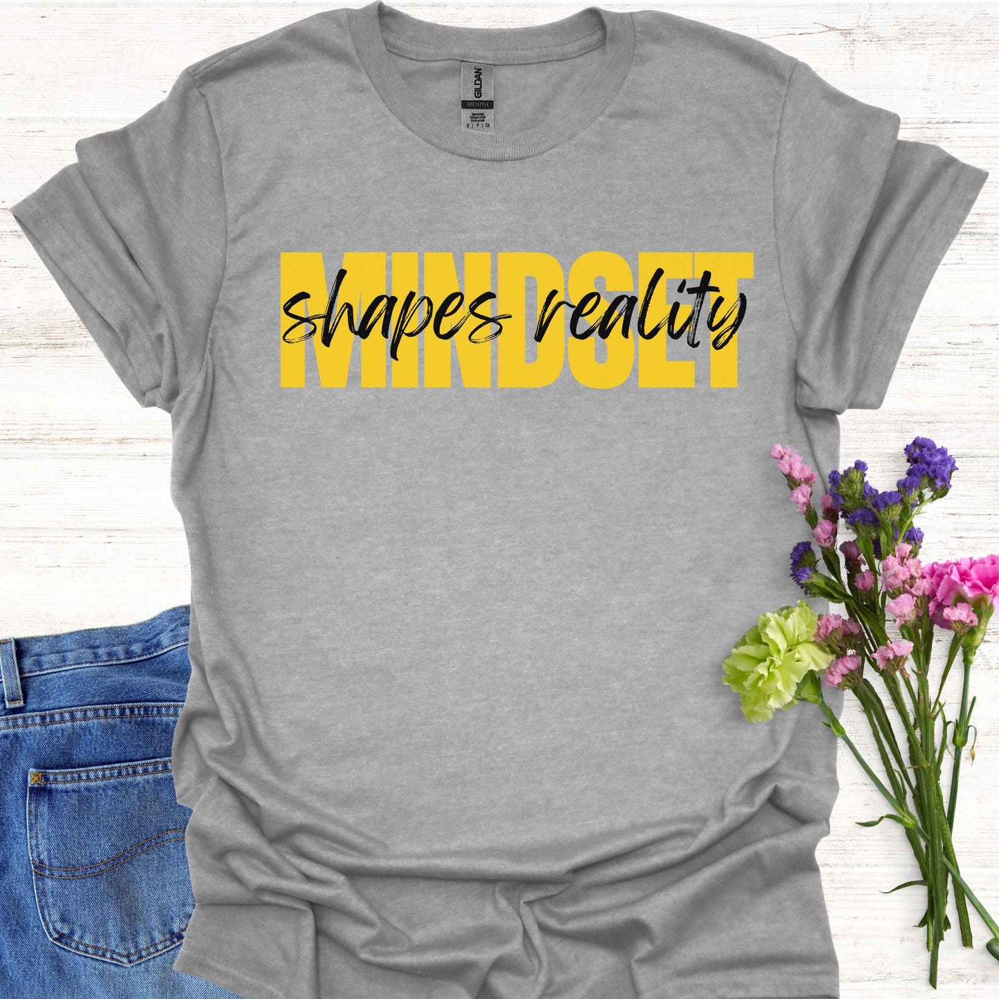 Grey t-shirt featuing a text-based design with the phrase 'Mindset Shapes Reality'. The word 'Mindset' is in bold yellow capital letters, while 'shapes reality' is in smaller cursive lettering. This design highlights how our mindset influences our perception and reality, empowering us to take control of our experiences and perspective in life.
