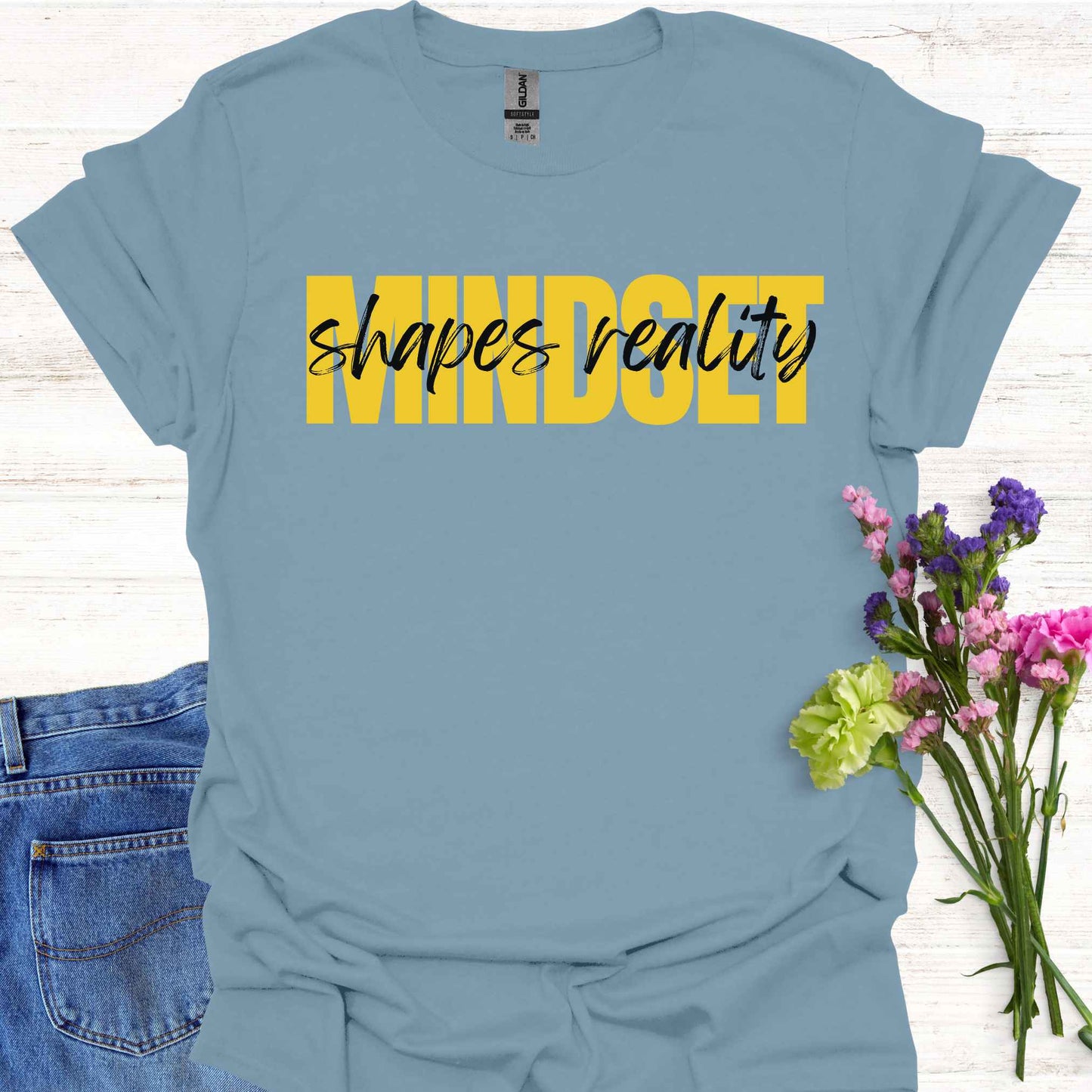 Blue t-shirt featuing a text-based design with the phrase 'Mindset Shapes Reality'. The word 'Mindset' is in bold yellow capital letters, while 'shapes reality' is in smaller cursive lettering. This design highlights how our mindset influences our perception and reality, empowering us to take control of our experiences and perspective in life.