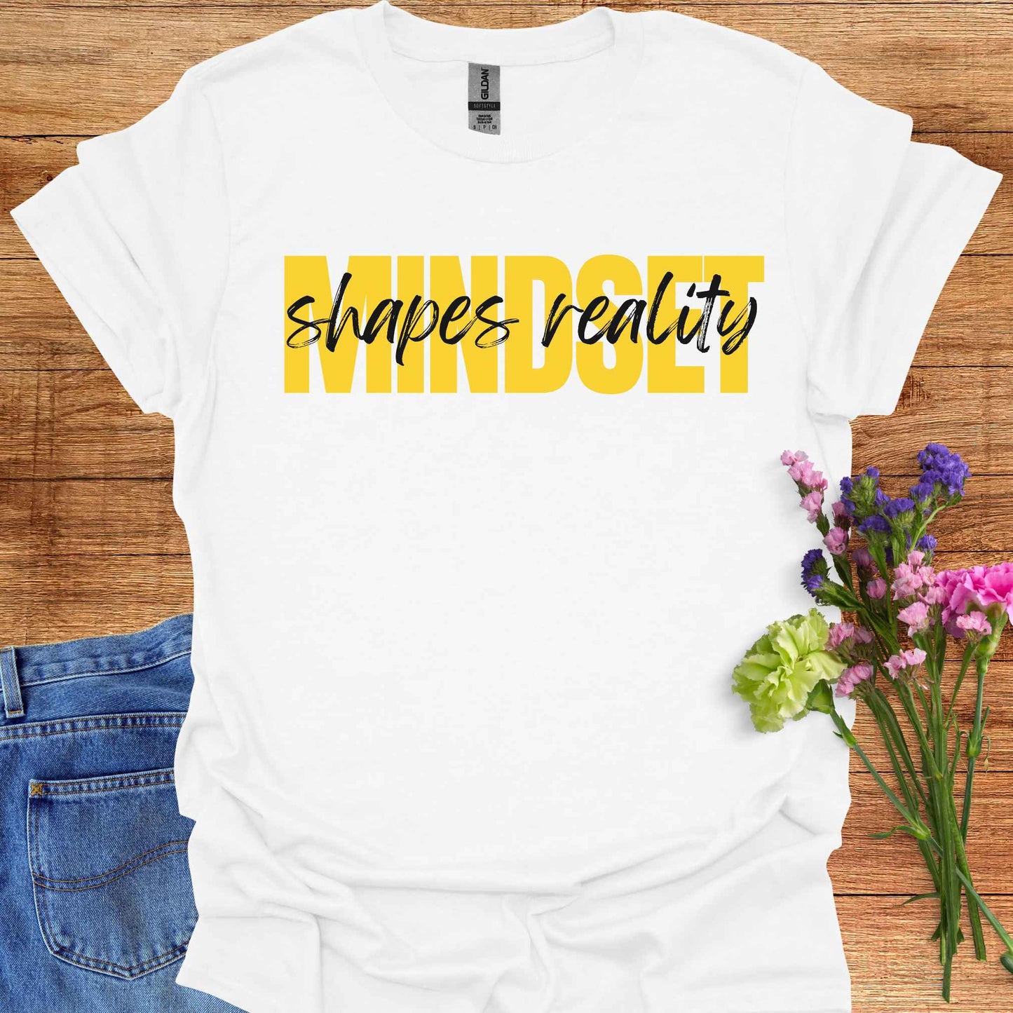 White t-shirt featuing a text-based design with the phrase 'Mindset Shapes Reality'. The word 'Mindset' is in bold yellow capital letters, while 'shapes reality' is in smaller cursive lettering. This design highlights how our mindset influences our perception and reality, empowering us to take control of our experiences and perspective in life.