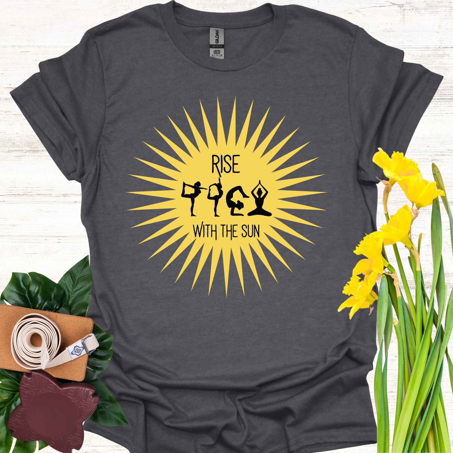 Dark grey t-shirt featuring a bright yellow sun graphic, with human forms in various yoga poses placed within the sun’s rays. The design is accompanied by the phrase 'Rise with the Sun,' symbolizing the connection between yoga, movement, and the natural rhythm of the day. This design encourages wearers to embrace vitality, inner balance, and strength, making it perfect for yoga practitioners and those seeking inspiration from both nature and mindful movement.