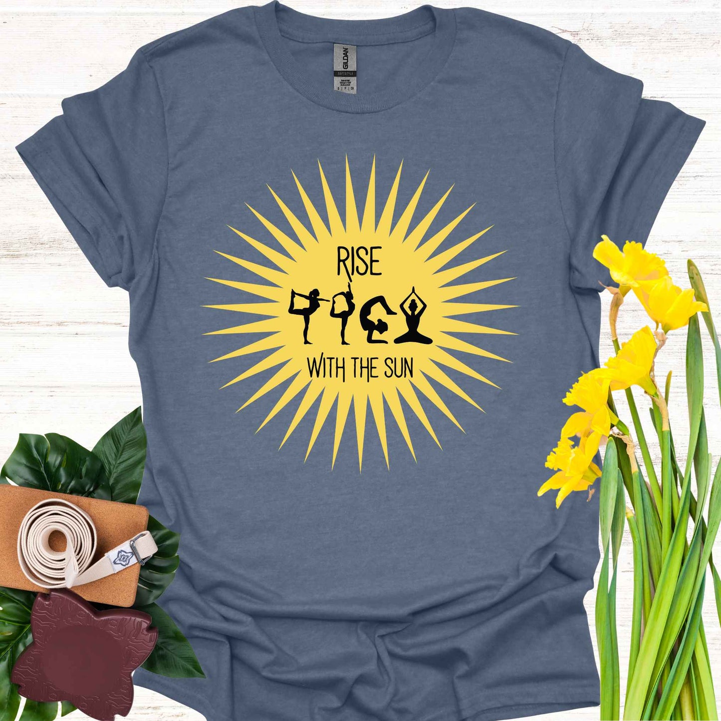 Blue t-shirt featuring a bright yellow sun graphic, with human forms in various yoga poses placed within the sun’s rays. The design is accompanied by the phrase 'Rise with the Sun,' symbolizing the connection between yoga, movement, and the natural rhythm of the day. This design encourages wearers to embrace vitality, inner balance, and strength, making it perfect for yoga practitioners and those seeking inspiration from both nature and mindful movement.