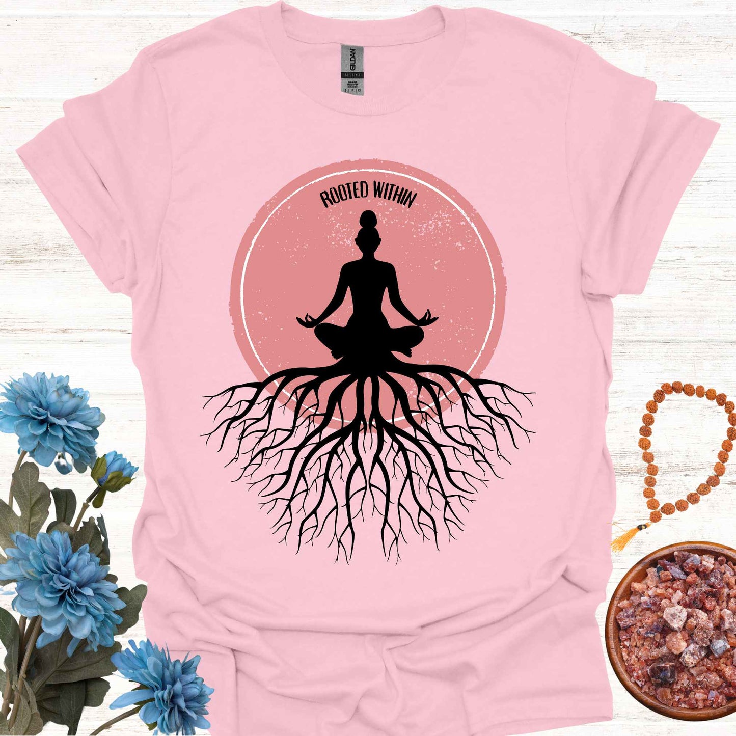 Light pink t-shirt featuring a graphic of deep tree roots extending downward, with the form of a meditating person sitting on top, suggesting that the roots belong to the meditator. This design symbolizes the yogic teaching of keeping awareness within, grounding oneself in inner peace, and embodying wisdom through meditation and spiritual practice