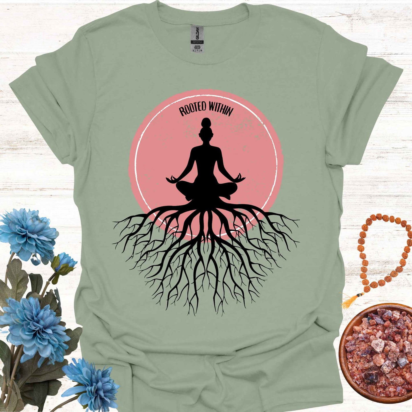 Sage green t-shirt featuring a graphic of deep tree roots extending downward, with the form of a meditating person sitting on top, suggesting that the roots belong to the meditator. This design symbolizes the yogic teaching of keeping awareness within, grounding oneself in inner peace, and embodying wisdom through meditation and spiritual practice.