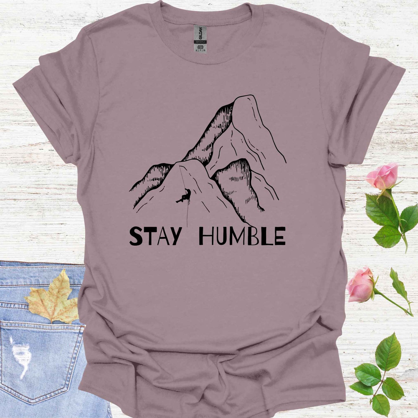 Light purple t-shirt featuring a graphic of a man climbing a vast mountain, appearing small and insignificant in comparison to the immense natural landscape. The phrase 'Stay Humble' accompanies the design, symbolizing the fragility of human life and the reminder of our humility in the face of nature's vastness and power.