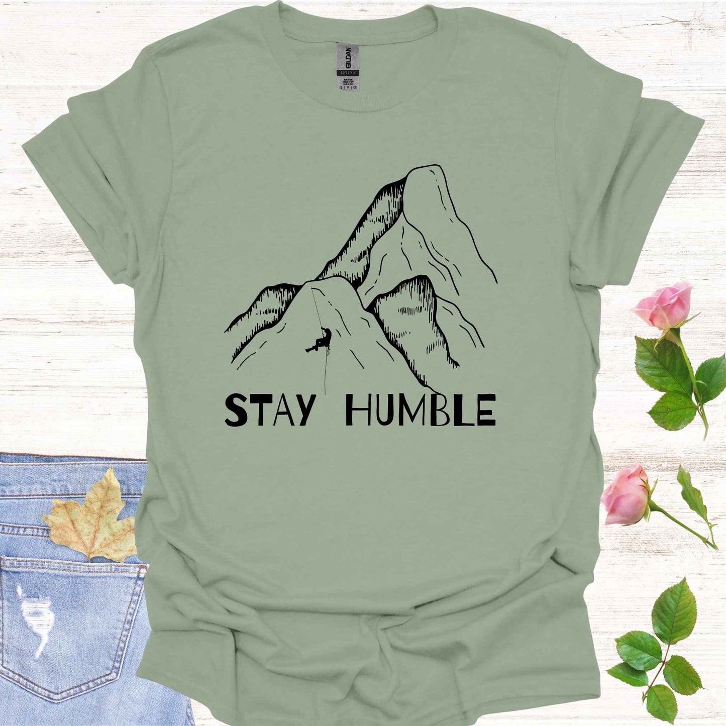 Sage green t-shirt featuring a graphic of a man climbing a vast mountain, appearing small and insignificant in comparison to the immense natural landscape. The phrase 'Stay Humble' accompanies the design, symbolizing the fragility of human life and the reminder of our humility in the face of nature's vastness and power.
