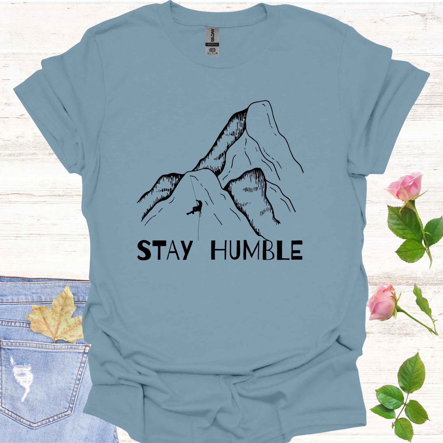 Blue t-shirt featuring a graphic of a man climbing a vast mountain, appearing small and insignificant in comparison to the immense natural landscape. The phrase 'Stay Humble' accompanies the design, symbolizing the fragility of human life and the reminder of our humility in the face of nature's vastness and power.