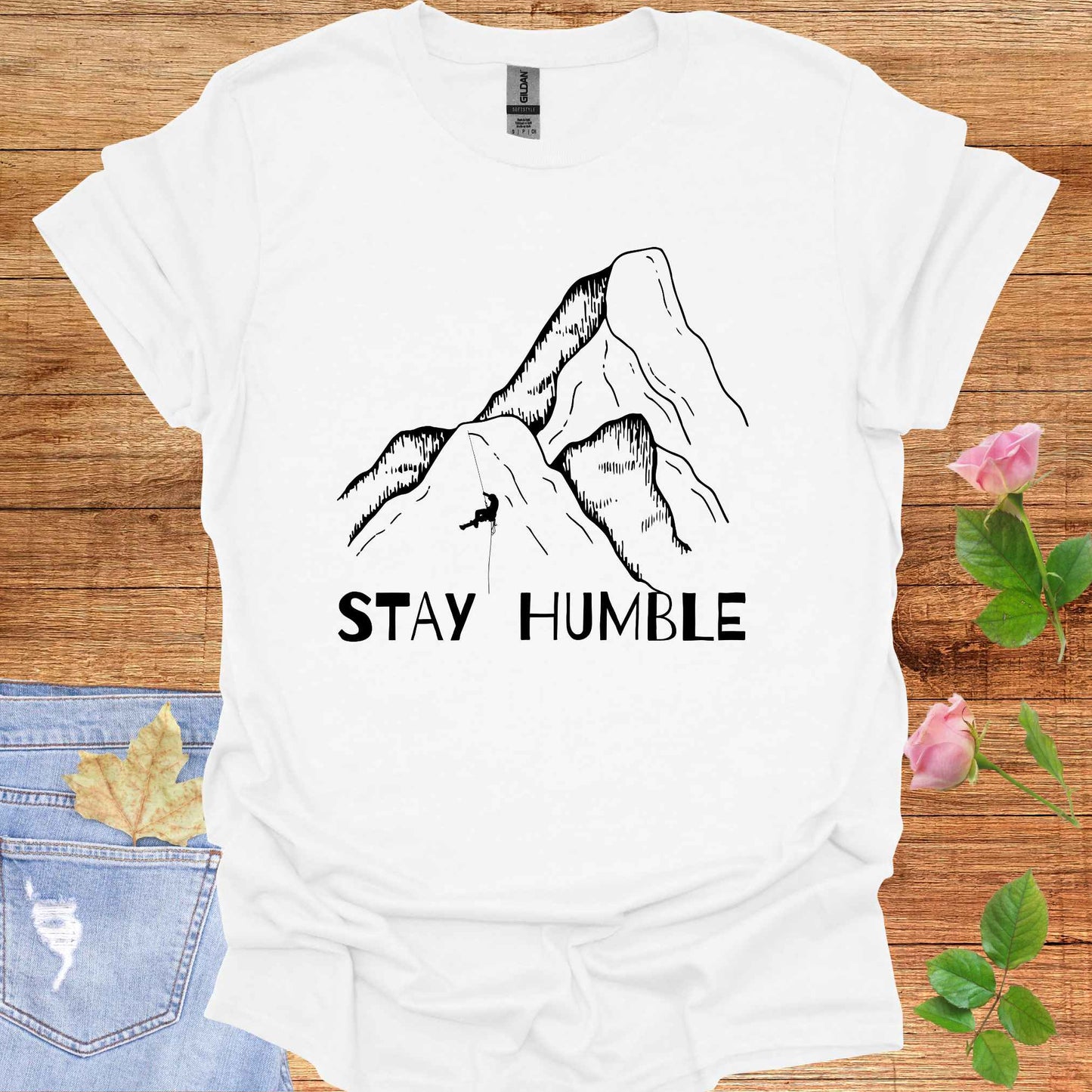 White t-shirt featuring a graphic of a man climbing a vast mountain, appearing small and insignificant in comparison to the immense natural landscape. The phrase 'Stay Humble' accompanies the design, symbolizing the fragility of human life and the reminder of our humility in the face of nature's vastness and power.