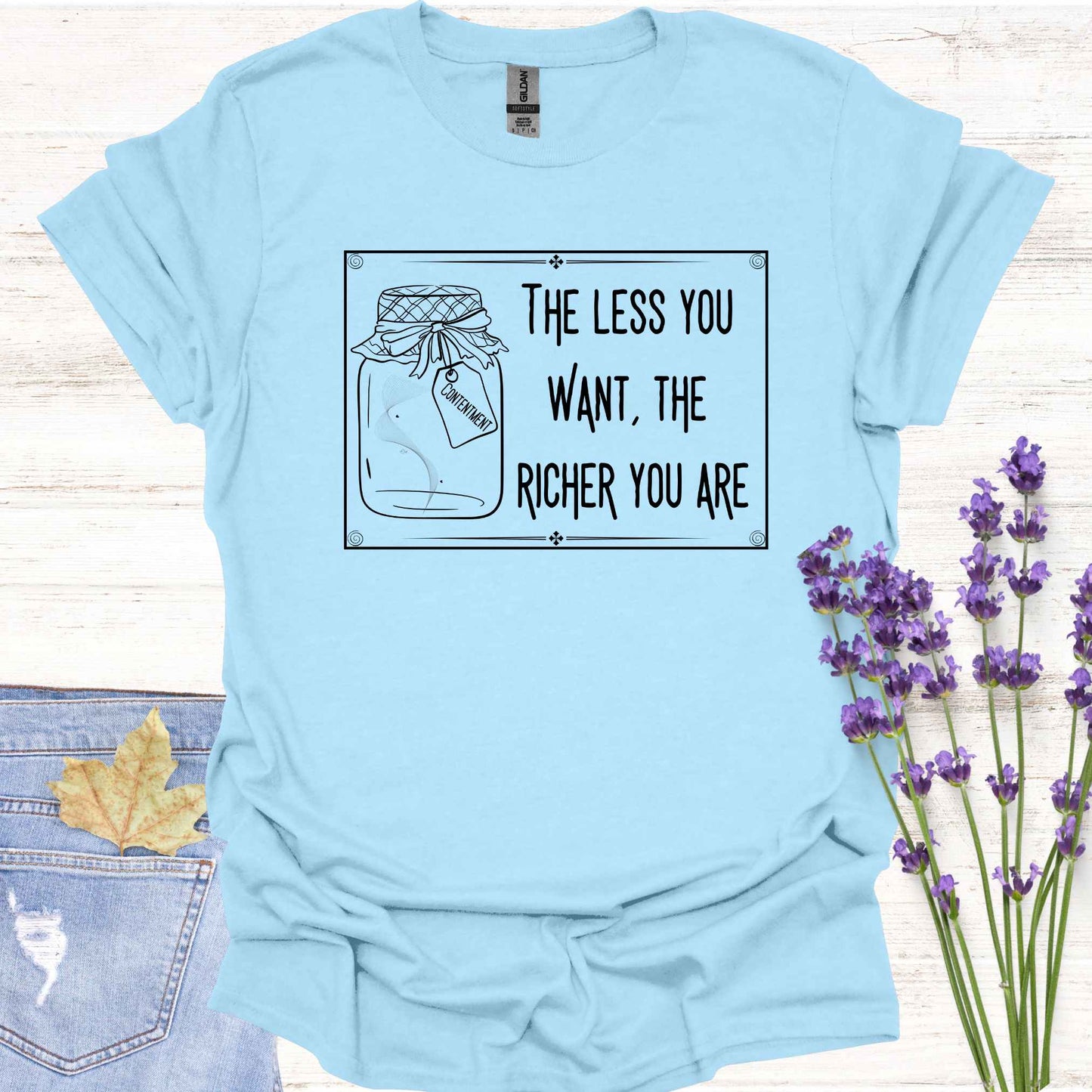 Light blue t-shirt featuring a framed black-and-white graphic of an empty jar labelled 'Contentment,' paired with the minimalist phrase 'The Less You Want, The Richer You Are.' This design symbolizes detachment, inner fulfillment, and the spiritual ideal of finding inner richness through desirelessness.