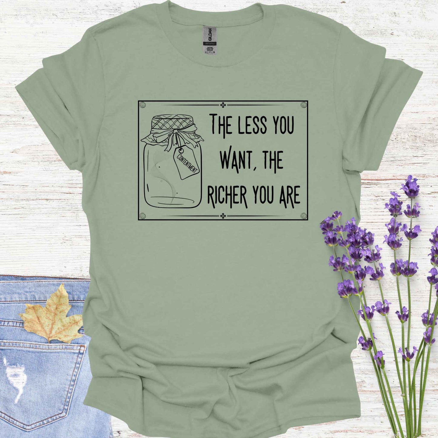 Sage green t-shirt featuring a framed black-and-white graphic of an empty jar labelled 'Contentment,' paired with the minimalist phrase 'The Less You Want, The Richer You Are.' This design symbolizes detachment, inner fulfillment, and the spiritual ideal of finding inner richness through desirelessness.