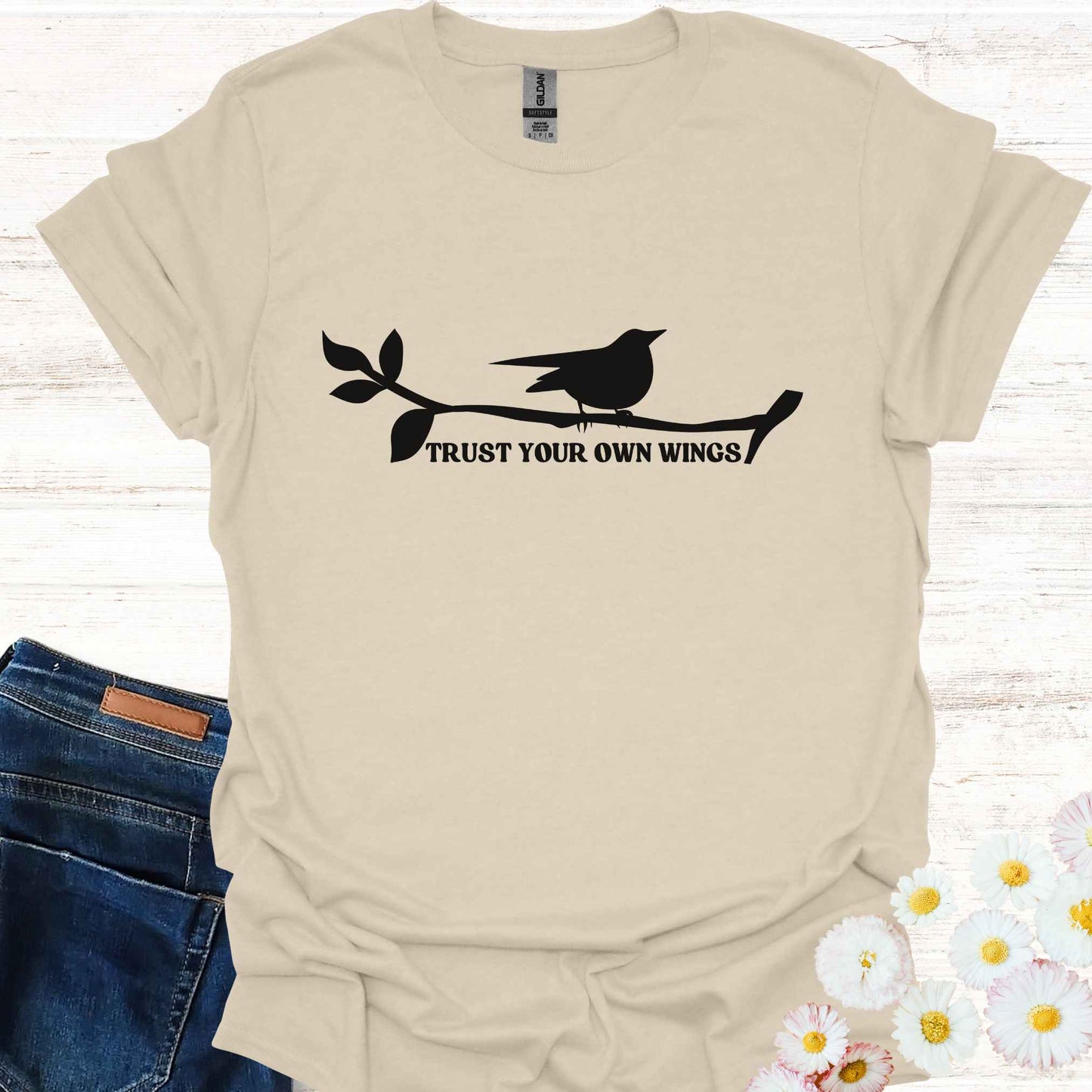 Beige t-shirt featuring a black graphic of a bird perched on a twig, with the empowering phrase 'Trust Your Own Wings.' This design symbolizes the spiritual teaching of self-reliance and trust, reminding us to have faith in our own abilities, just as a bird trusts its wings rather than the branch beneath it.
