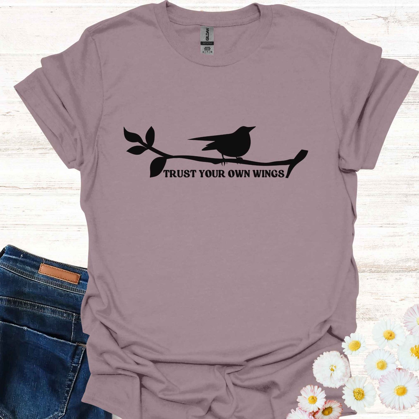 Light purple t-shirt featuring a black graphic of a bird perched on a twig, with the empowering phrase 'Trust Your Own Wings.' This design symbolizes the spiritual teaching of self-reliance and trust, reminding us to have faith in our own abilities, just as a bird trusts its wings rather than the branch beneath it.