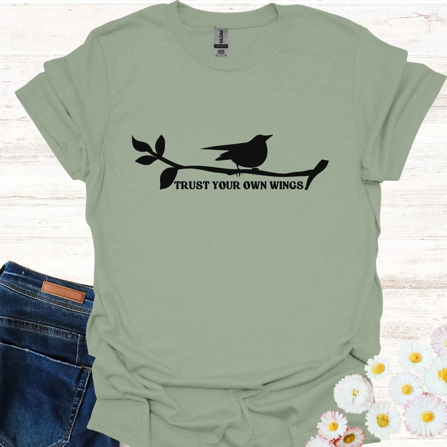Sage green t-shirt featuring a black graphic of a bird perched on a twig, with the empowering phrase 'Trust Your Own Wings.' This design symbolizes the spiritual teaching of self-reliance and trust, reminding us to have faith in our own abilities, just as a bird trusts its wings rather than the branch beneath it.