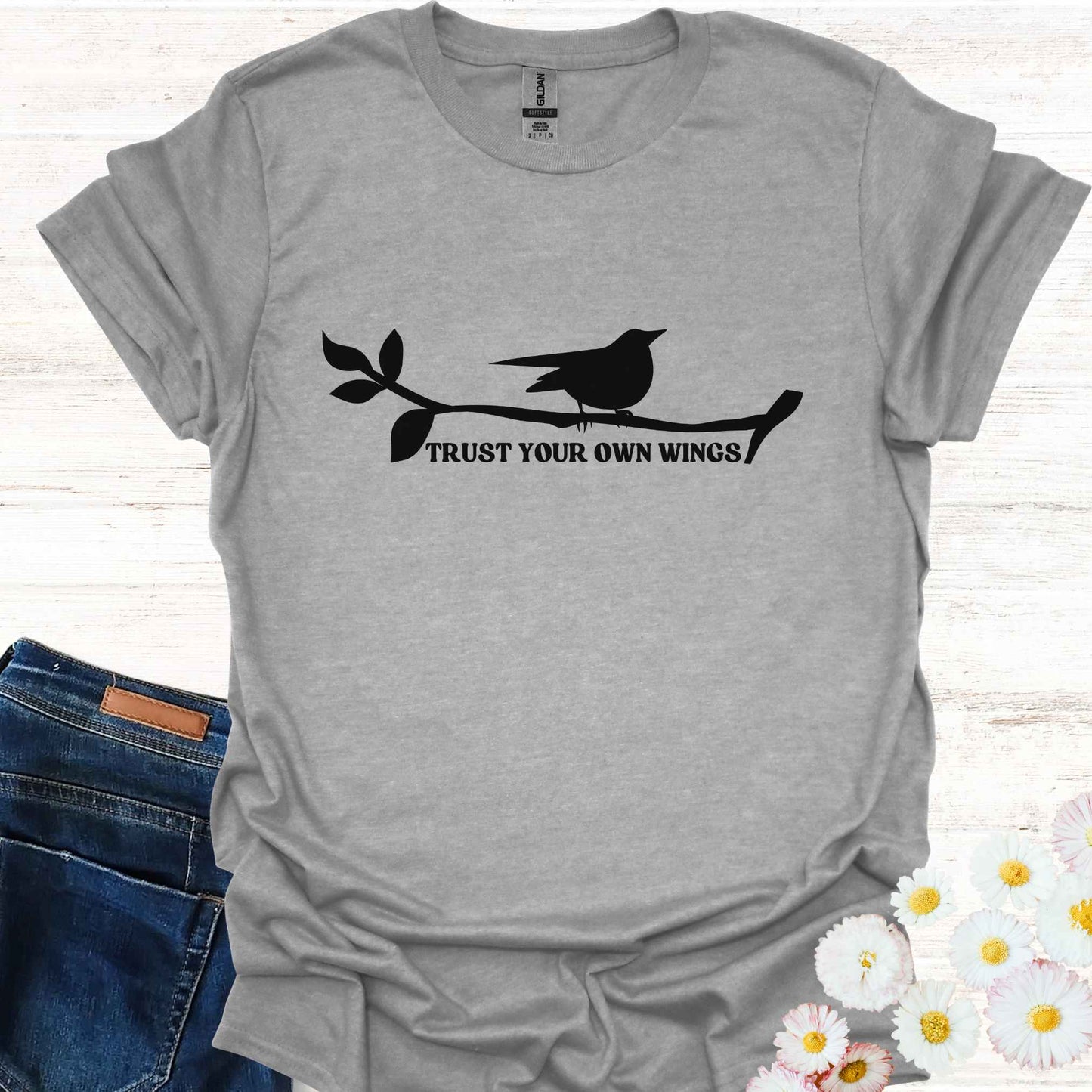 Grey t-shirt featuring a black graphic of a bird perched on a twig, with the empowering phrase 'Trust Your Own Wings.' This design symbolizes the spiritual teaching of self-reliance and trust, reminding us to have faith in our own abilities, just as a bird trusts its wings rather than the branch beneath it.