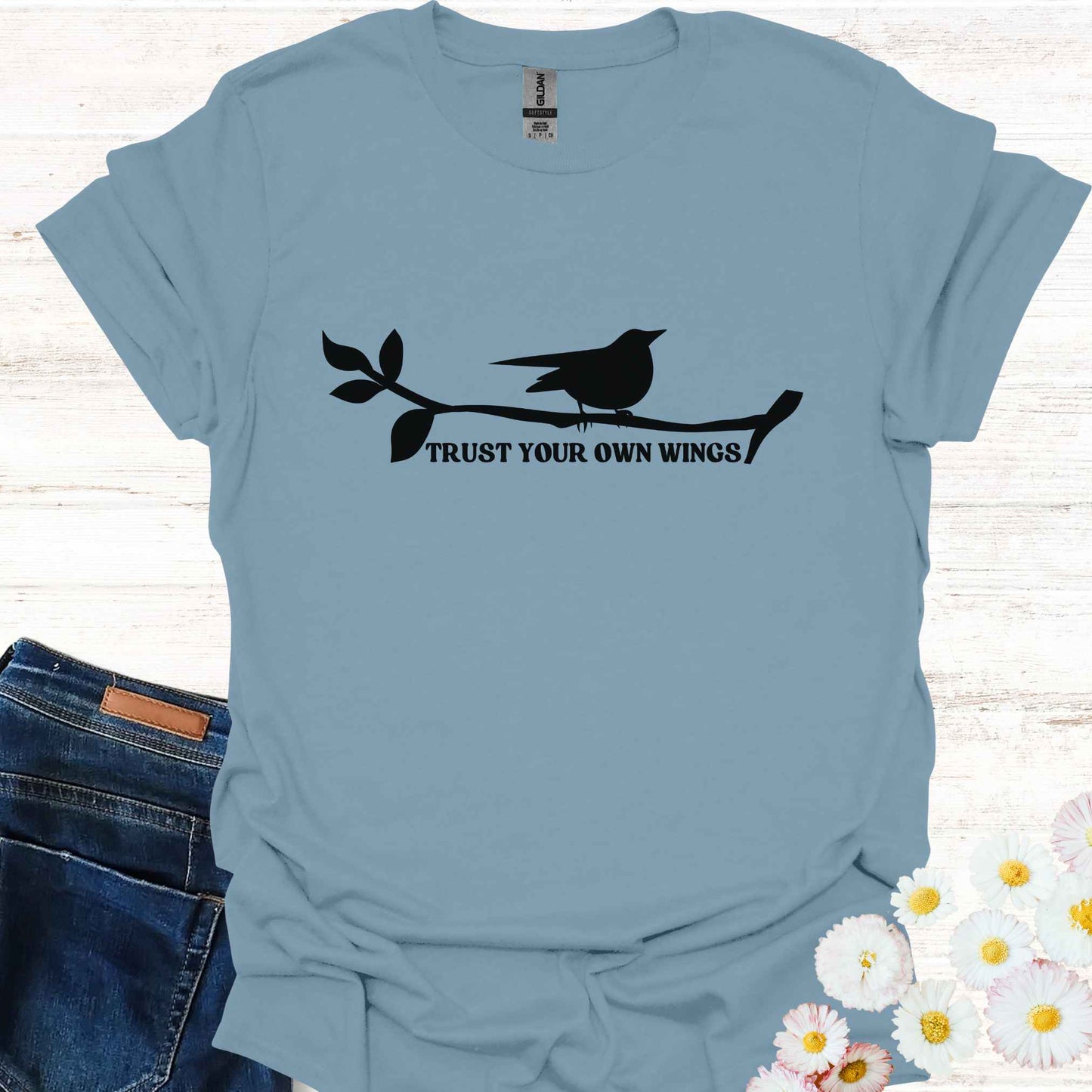 Blue t-shirt featuring a black graphic of a bird perched on a twig, with the empowering phrase 'Trust Your Own Wings.' This design symbolizes the spiritual teaching of self-reliance and trust, reminding us to have faith in our own abilities, just as a bird trusts its wings rather than the branch beneath it.
