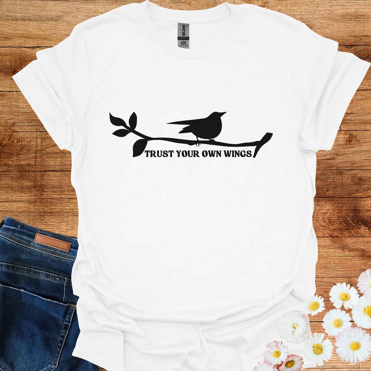 White t-shirt featuring a black graphic of a bird perched on a twig, with the empowering phrase 'Trust Your Own Wings.' This design symbolizes the spiritual teaching of self-reliance and trust, reminding us to have faith in our own abilities, just as a bird trusts its wings rather than the branch beneath it.