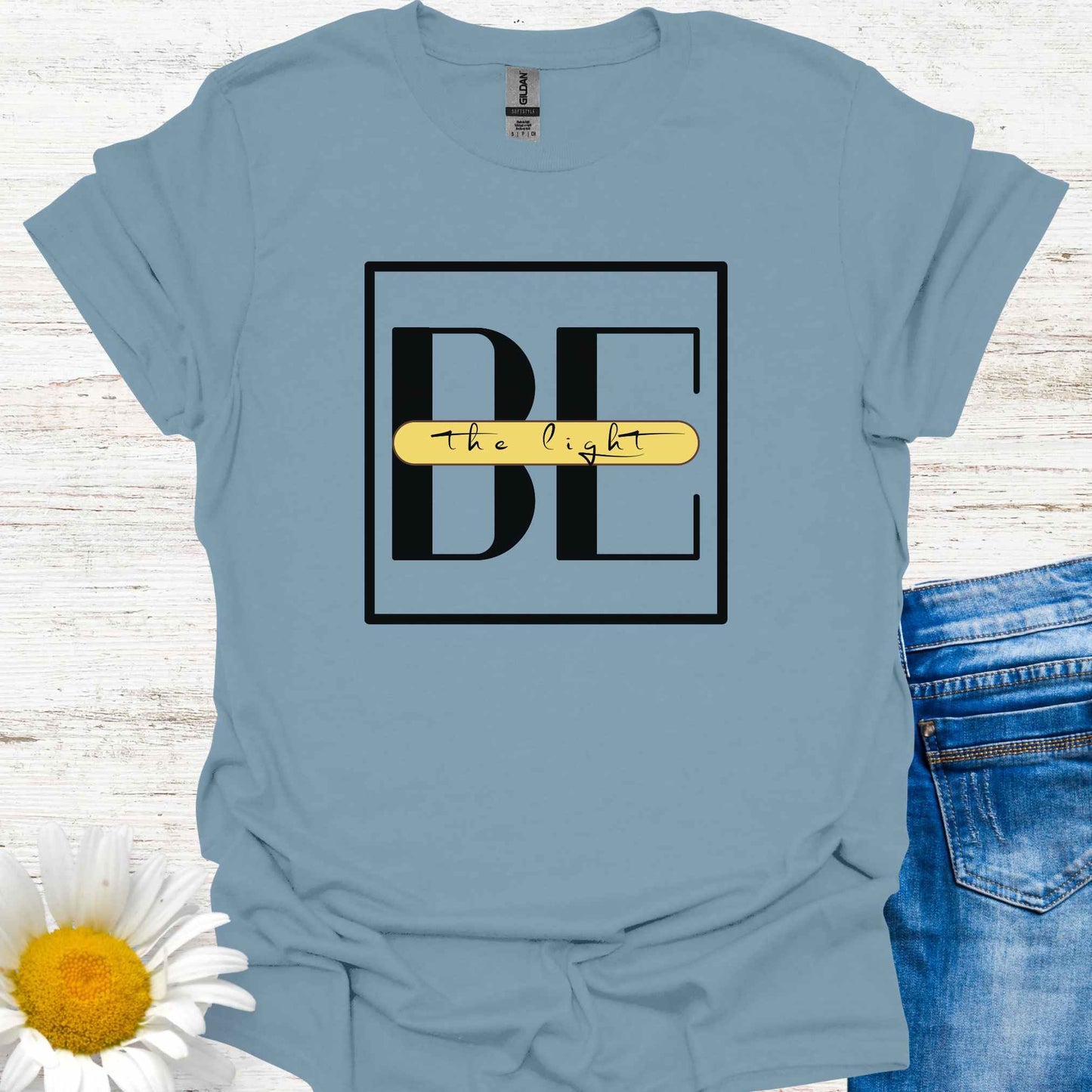 Blue t-shirt featuring the phrase 'Be the Light,' with the word 'BE' in large letters and the small cursive words 'the light' written across it on a yellow banner. This design encourages positivity, inspiration, and personal empowerment, reminding wearers to shine their light and spread kindness in the world.